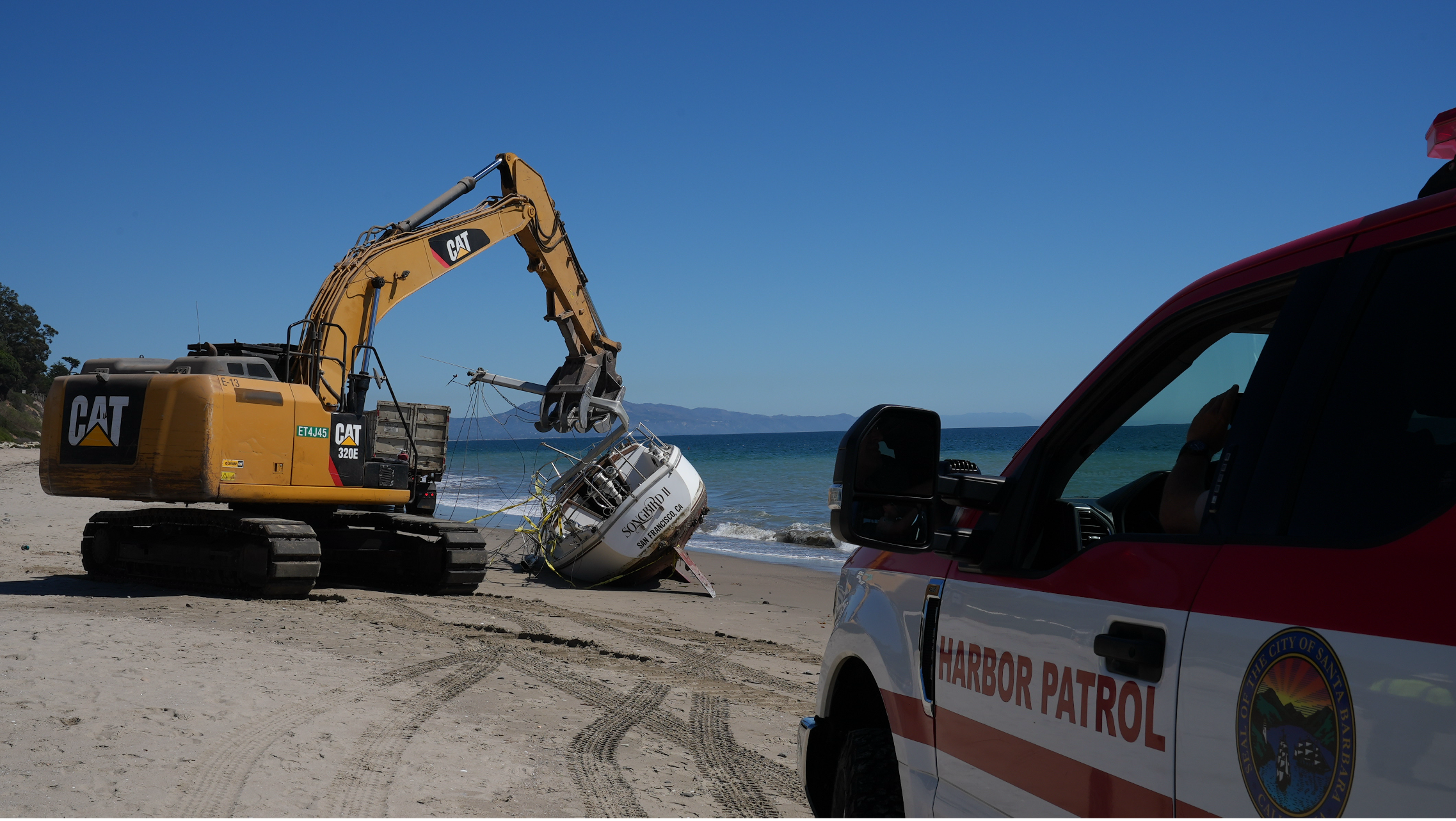 A Harbor Patrol vehicle and a piece of salvage equipment on the beach near a derelict boat.