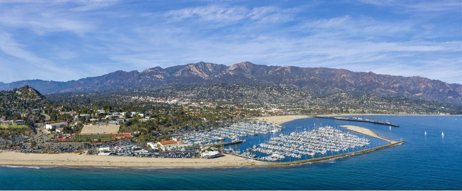 A birds eye view of Santa Barbara with boats in blue ocean at the harbor and mountains in the distance.