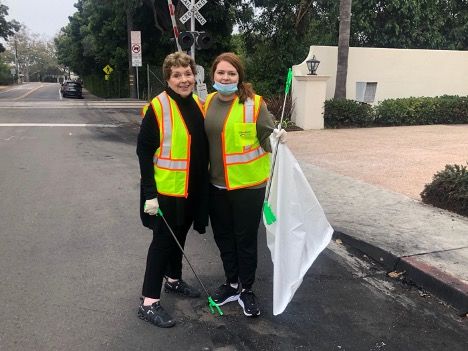 Two female Montecito residents wearing yellow vests carrying a white plastic trash bag and long metal tongs to pick up trash