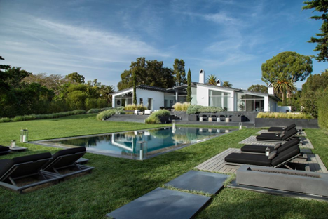 Large contemporary Santa Barbara home, white and glass, beyond a pool and sprawling lawn, and under a blue sky.