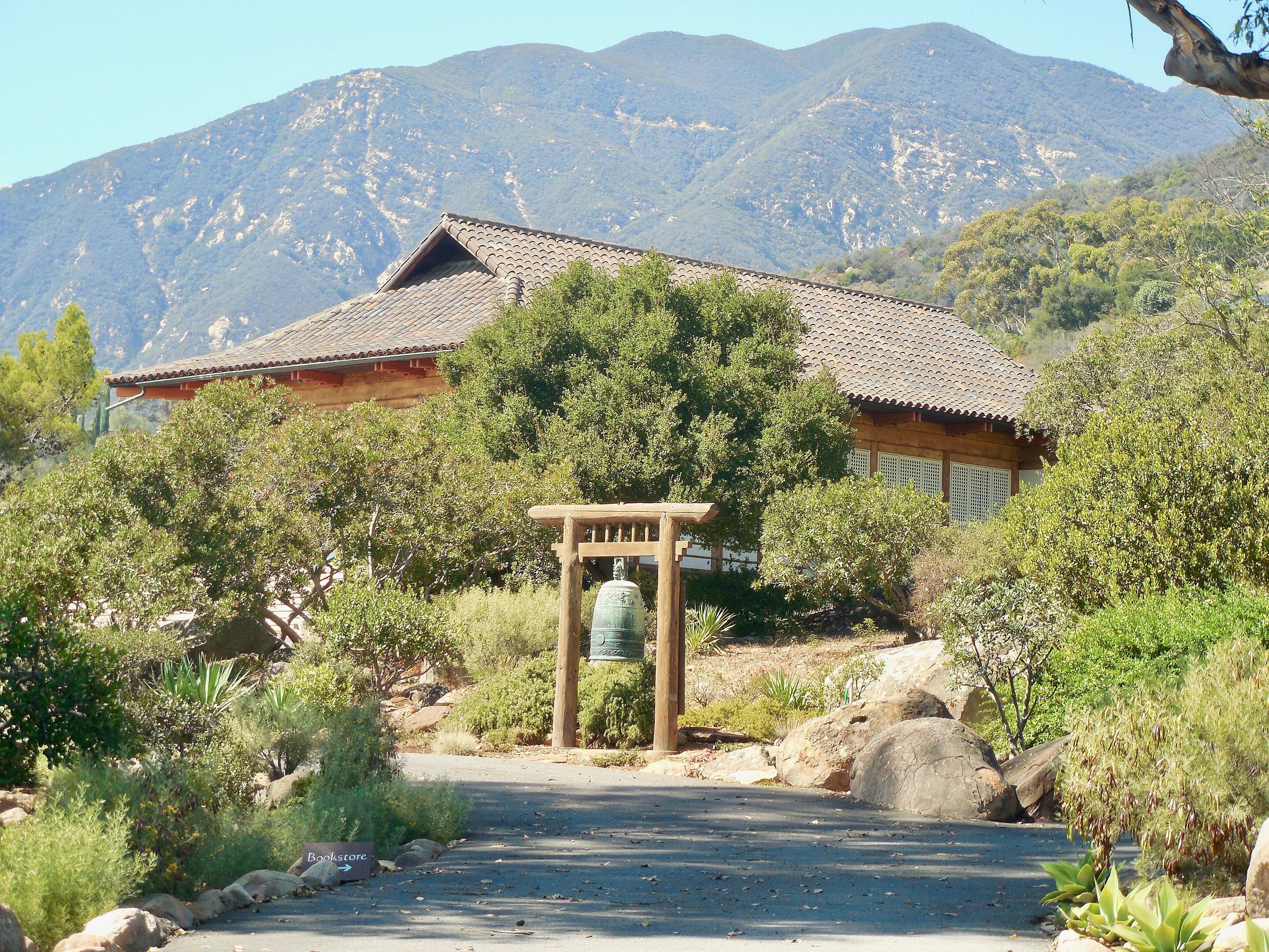 A large bell hanging at the front of a temple in Santa Barbara with green foliage in front and the mountains in the background.