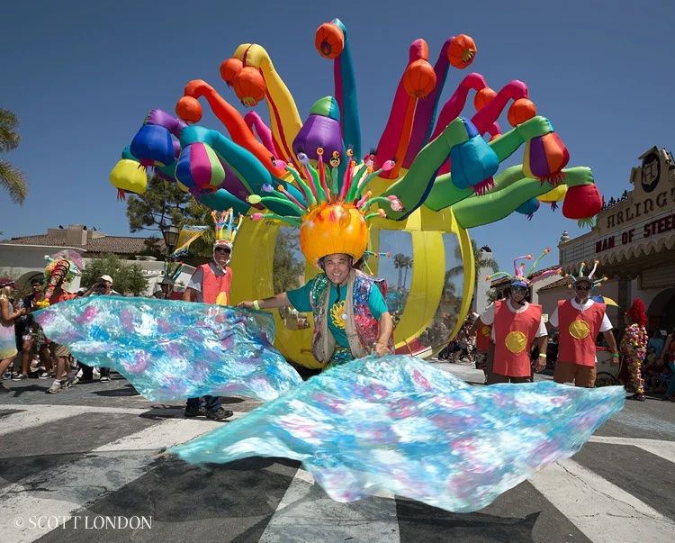 A magical float in the Santa Barbara Solstice parade, with a man wearing a wild and colorfully decorated hat in the center surrounded by tall balloons and a shiny piece of fabric in front of him