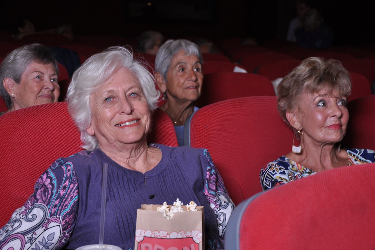 Four senior women smiling in red seats inside a movie theater. The one in the front looking at the camera is holding a bag of popcorn