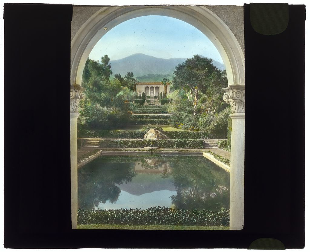 Vintage photo of an early Spanish Revival-style Montecito mansion, with a pool and lush gardens in front and mountains in the background