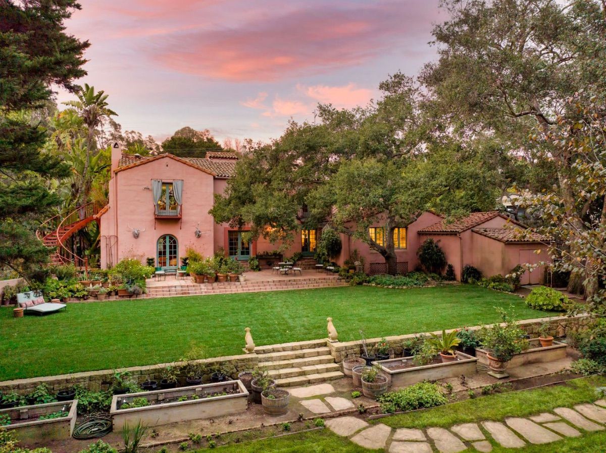 A romantic, coral tinted Montecito home with a sunset sky above, surrounded by lush trees and grass with an outside staircase on the site and a front year filled with grass and a stone trail