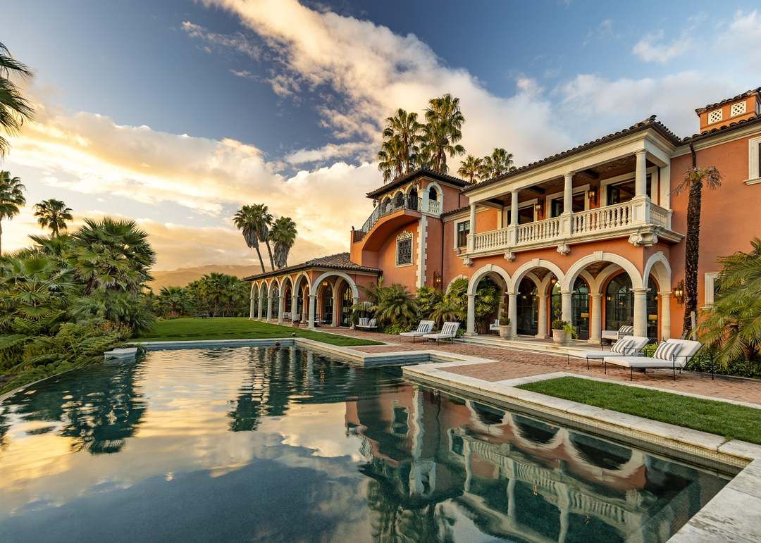 An ochre-colored luxury Hacienda-style Spanish Colonial estate with a refreshing pool in the foreground and lots of lush landscaping.
