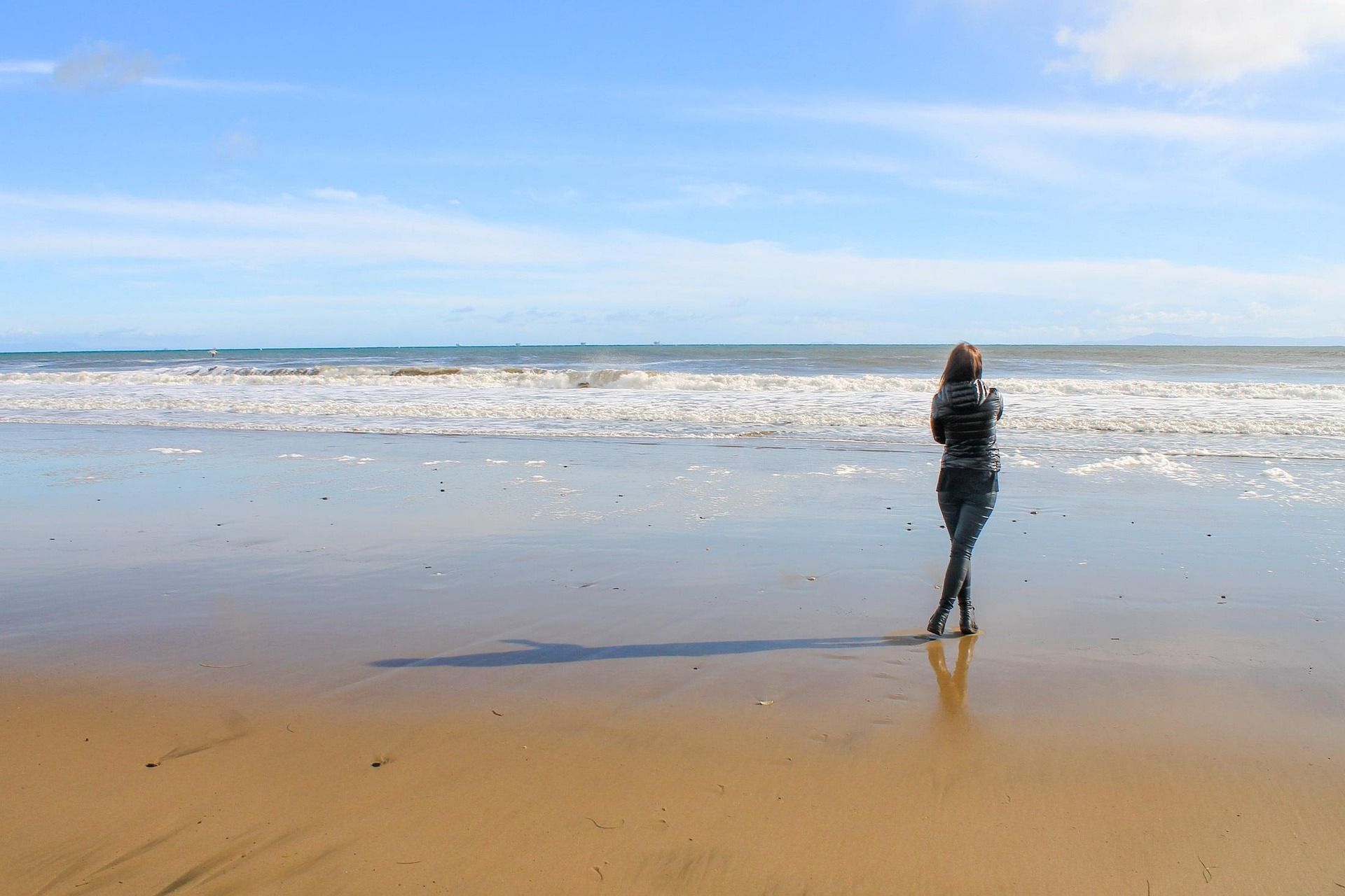 A woman standing on the sand of a Santa Barbara beach watching the gentle waves roll in against a blue sky.