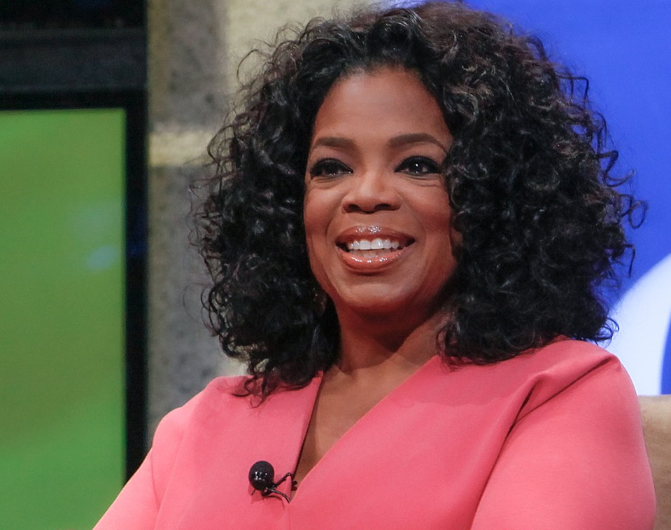 Oprah Winfrey, with a big smile sitting in a pink top with a microphone clipped to it