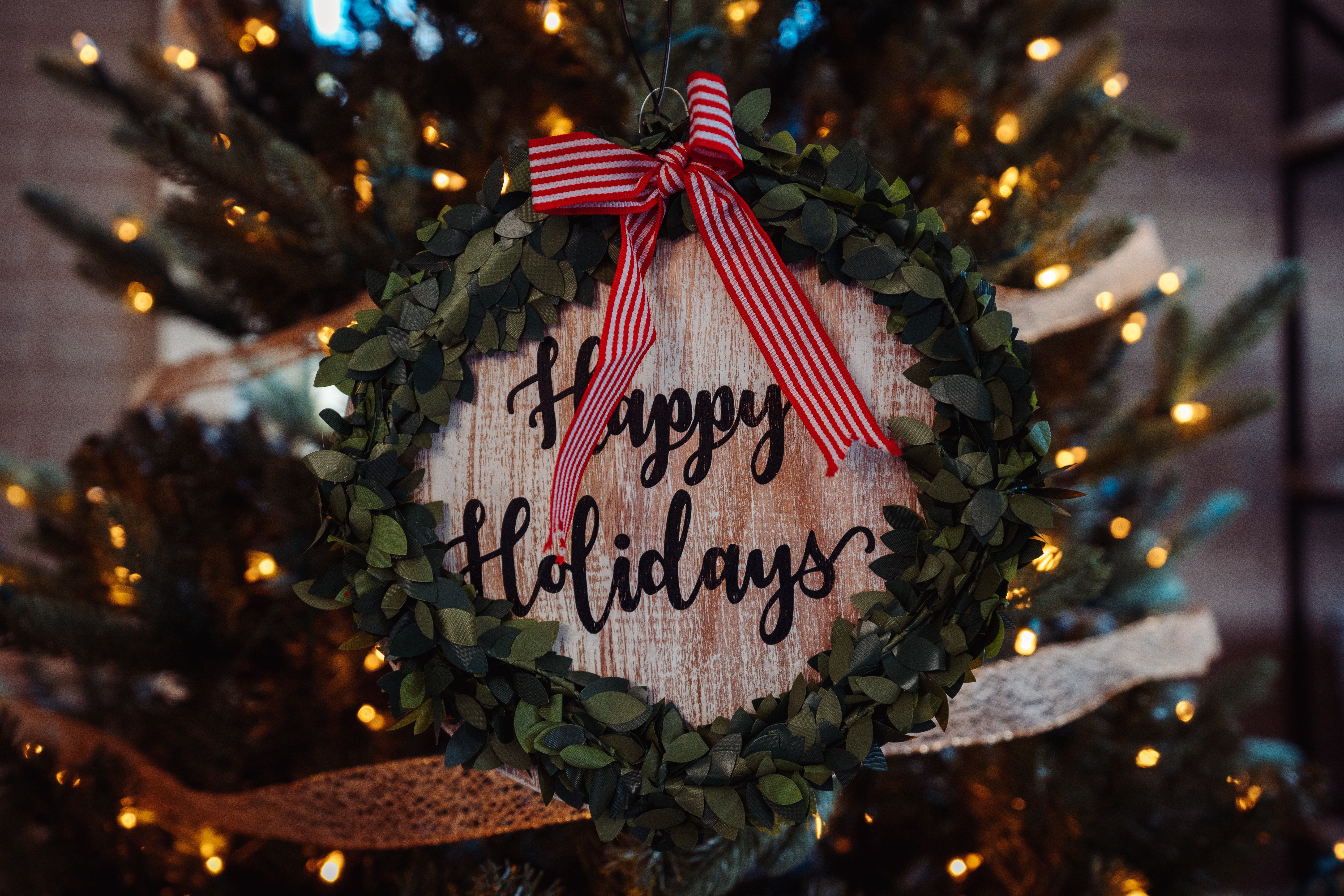 A round piece of wood with the words Happy Holidays carved into it with lights, greens and a big red bow on top