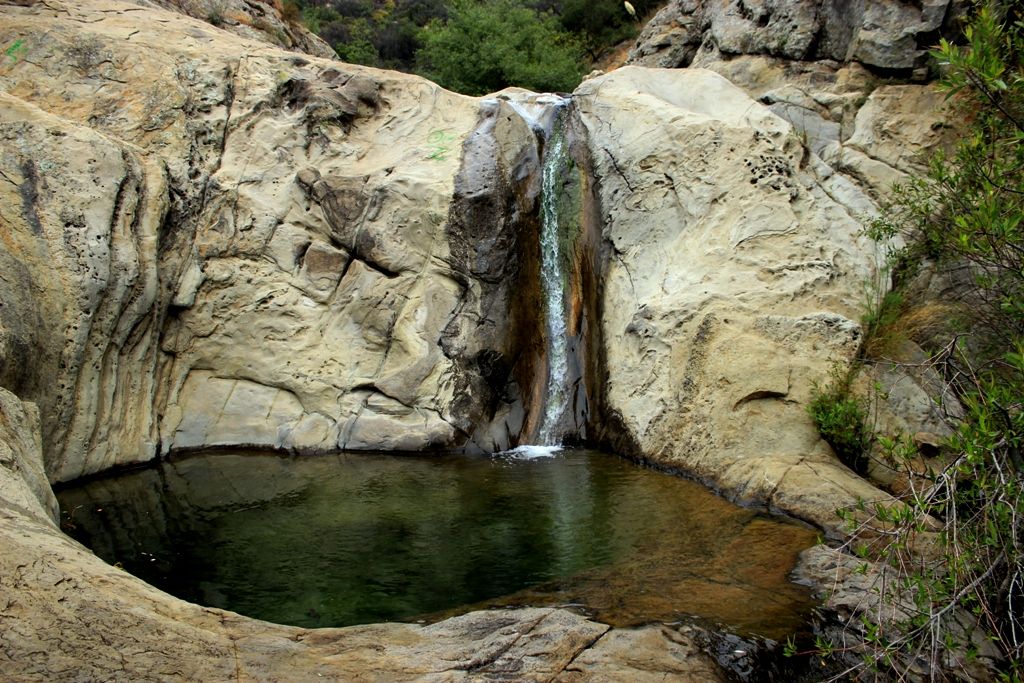 A small, delightful waterfall over a rock wall maybe 10 feet high, cascading into a clear rock basin.