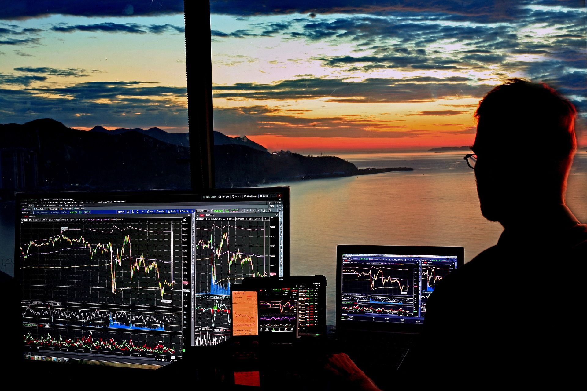 A man sitting at dusk in front of several computer monitors with the sunset in the background.
