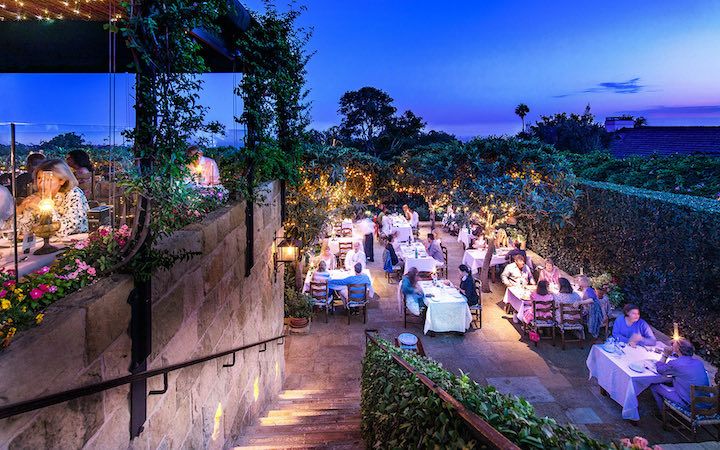 Aerial view of the romantic outside patio of the Stonehouse Restaurant at San Ysidro Ranch at dusk with a deep blue sky and ocean in the background.