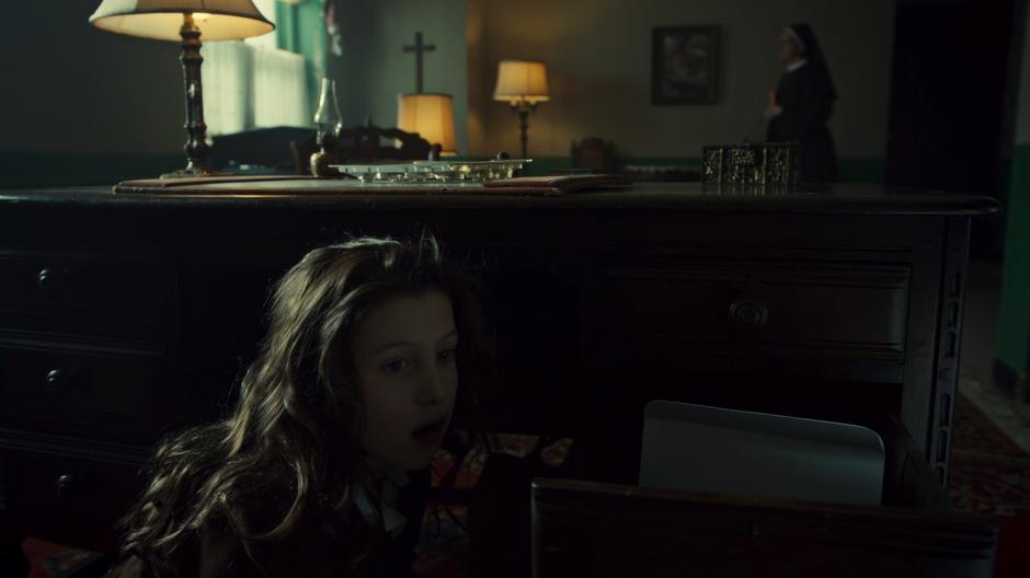 Helena hides behind the desk eating chocolates while one of the nuns enters the room and heads to the couch.