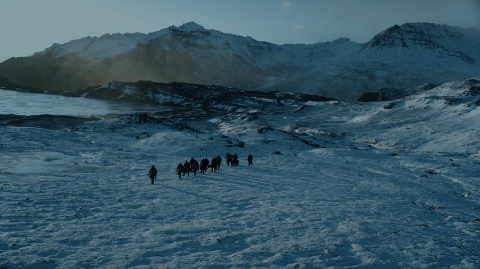 The party walks through the snow on the hillside before some mountains.