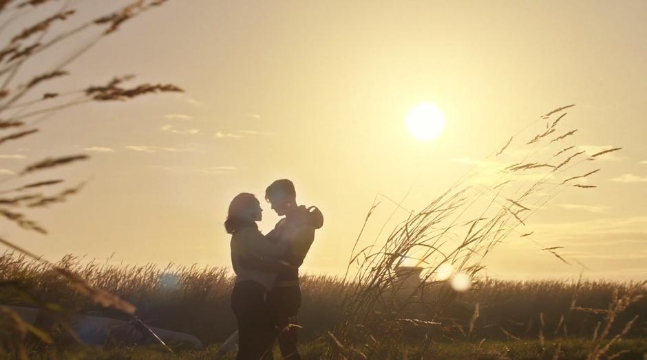 Iris and Barry embrace in the field in front of the setting sun.