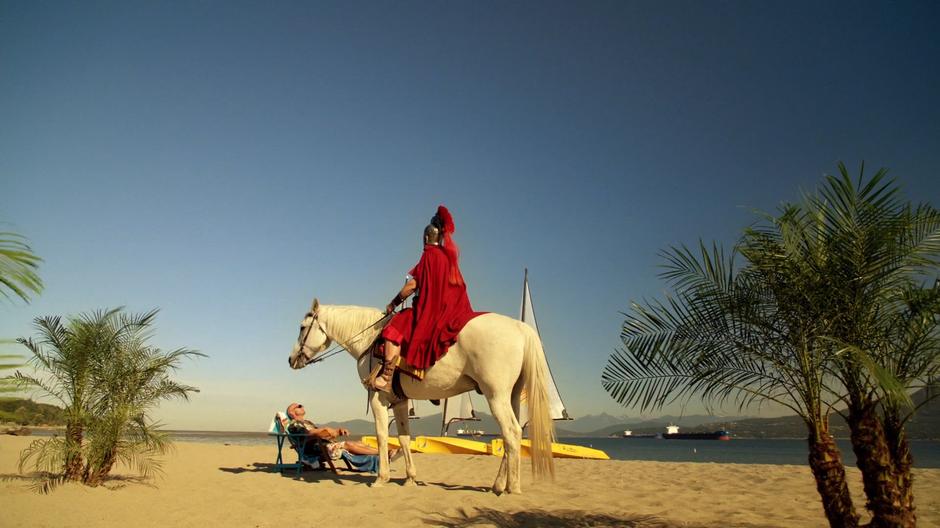 Julias Caesar sits on his horse which is blocking the sun from Mick who is lounging on the beach.