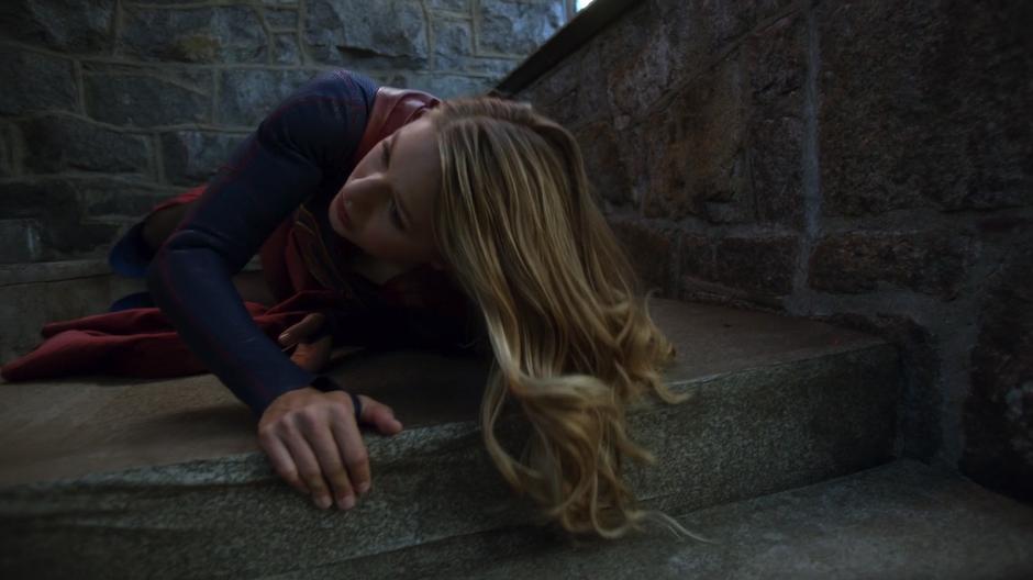 Kara struggles to sit up on the steps beside the building.