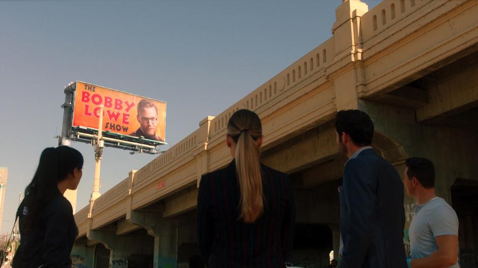 Ella, Chloe, Lucifer, and Dan look up at the Bobby Lowe Show billboard.