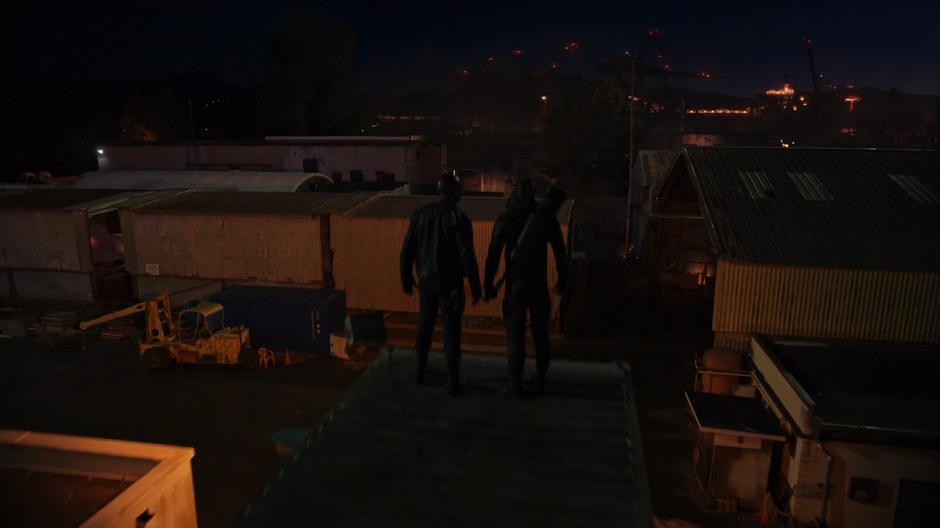 Diggle and Oliver stand atop a shipping container and survey the dockyard.