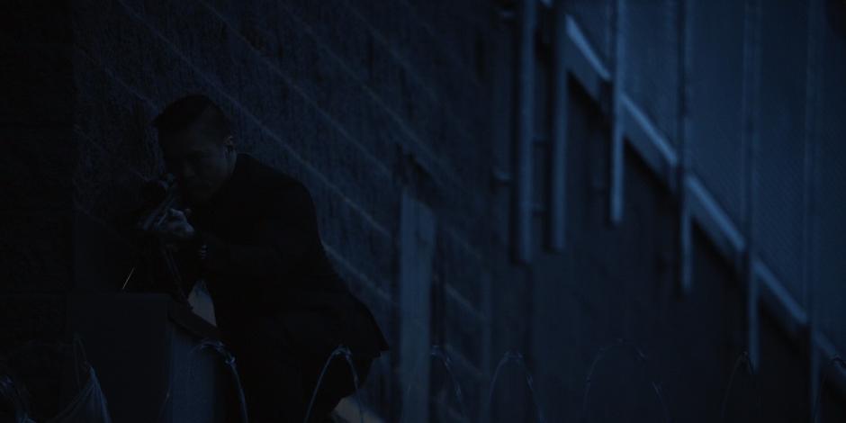 A mercenary settles at the edge of the roof with a rifle looking down at the base.