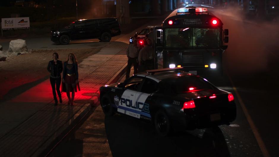 Alex and Kara walk away from the transport which is being secured by the police.