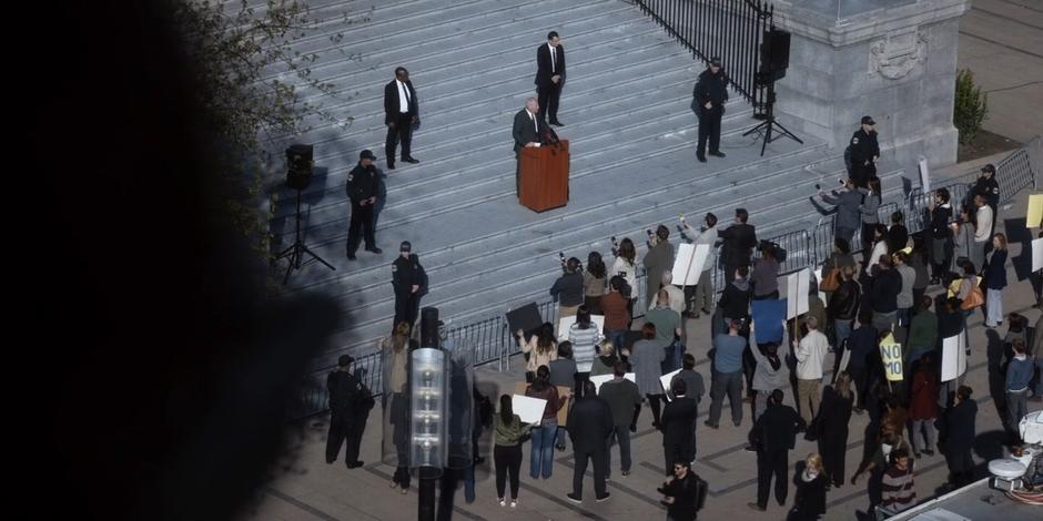 Ted Bishop speeds at a lectern on the steps of the building in front of a crowd while Carly aims at him from the nearby rooftop.