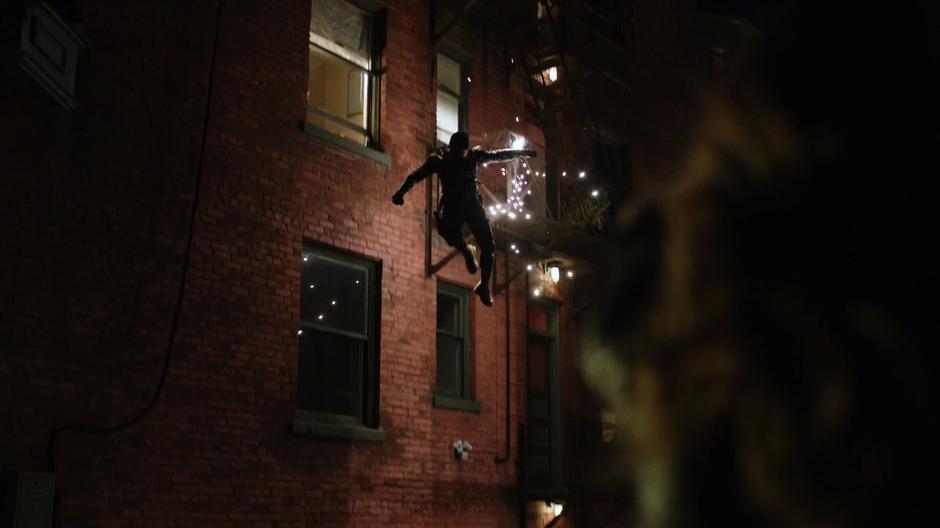 The Vigilante jumps down from the fire escape as Dinah shoots at him.