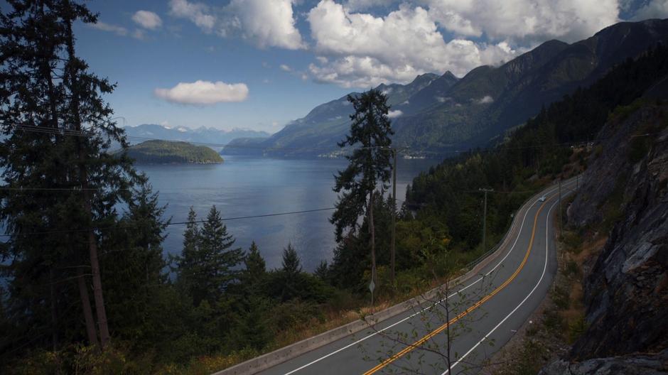 Kara drives Alex down a highway overlooking the water and mountains.