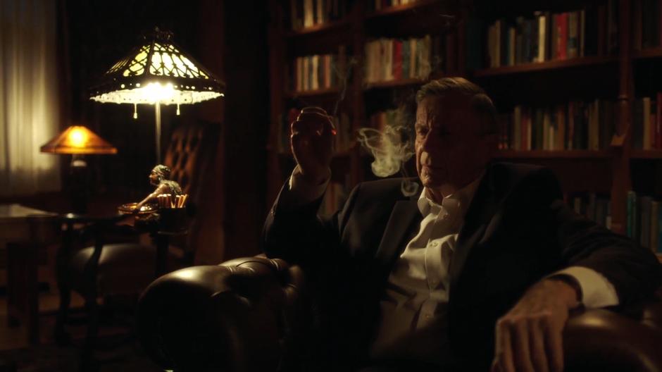 The Smoking Man sits in his chair in the lounge.