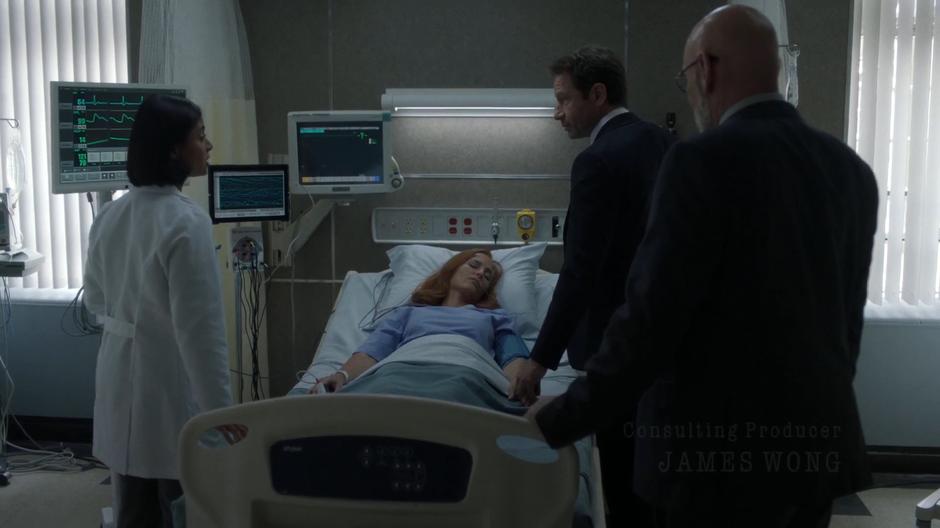 Dr. Joyet talks to Mulder and Skinner over Scully's hospital bed.