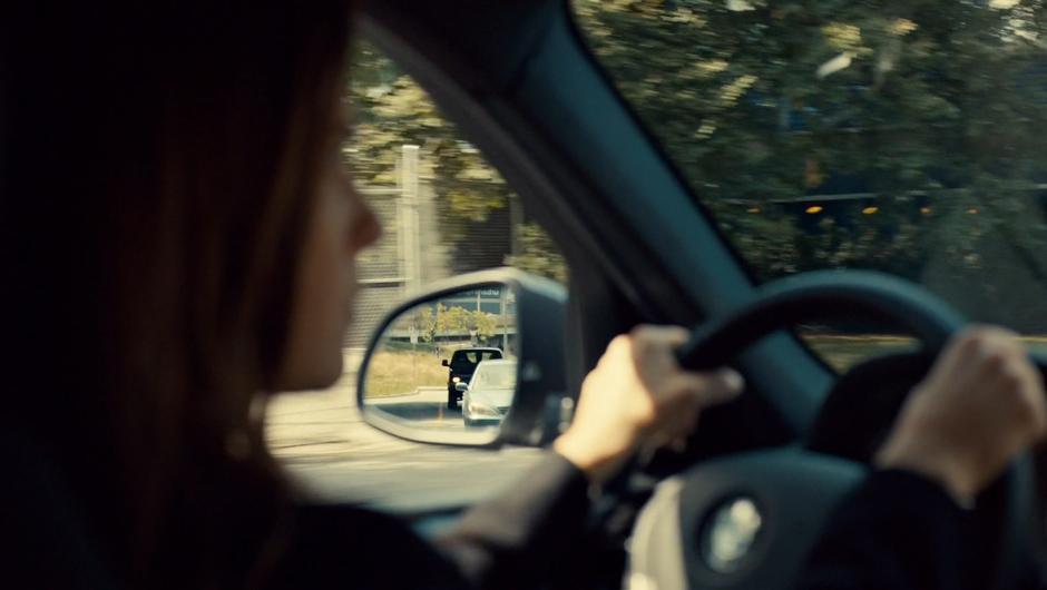 Mary drives down the road while a black van is visible in her side view mirror.