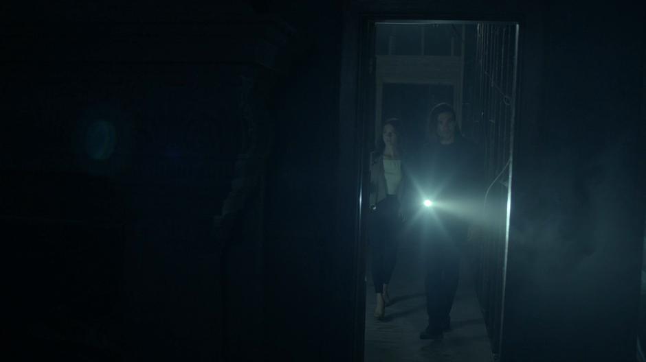 Julia and Quentin walk in through the entrance hallway into a darkened room.