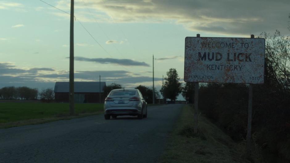 Mulder and Scully drive past the Welcome to Mud Lick sign.