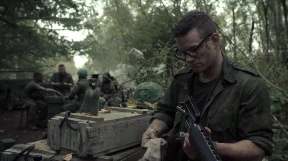 Skinner cleans his rifle while John James talks about his ear collection to a group of soldiers.