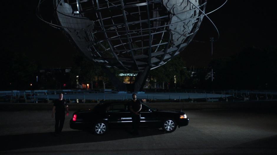 Two goons stand outside a black car waiting in front of the Unisphere.