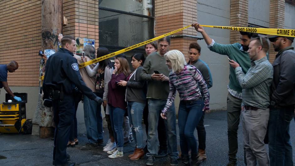 Ravi holds up the police tape for Liv as an officer keeps the crowd in check.