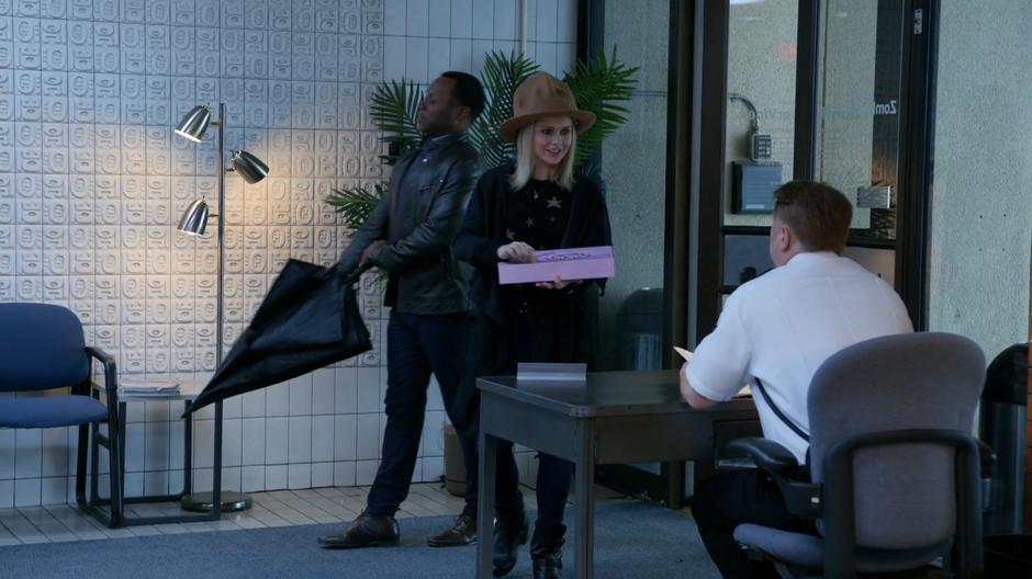 Clive puts away his umbrella while Liv holds out the box of donuts to the security guard.