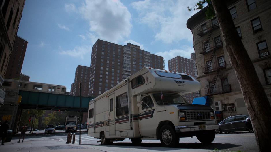 The RV sits parked on the side of the road while a subway drives past on the elevated rails.