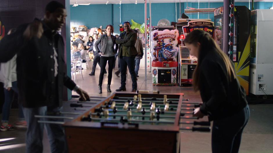 Alex and J'onn watch happily as M'yrnn and Ruby play a game of foosball.