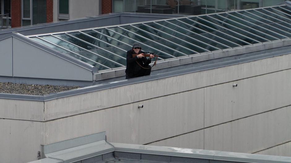 The killer looks over from the adjacent rooftop at Alex while holding a rifle.