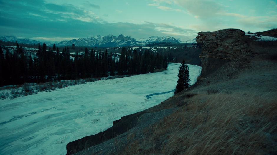 A frozen river runs beneath the cliff as the snowy mountains sit in the background.