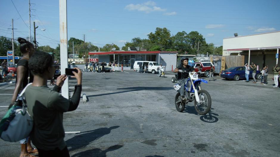 People watch a guy does motorcycle tricks in the parking lot.