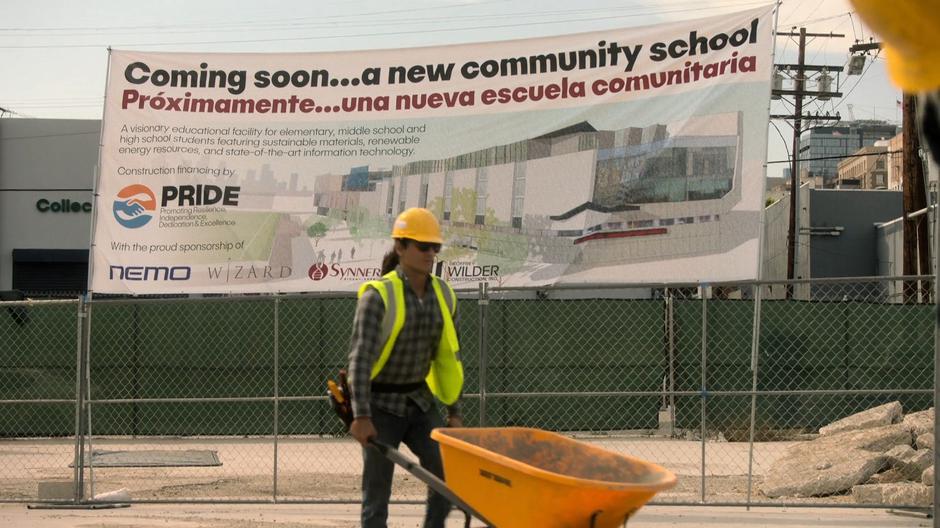 A construction worker pushes a wheelbarrow in front of a sign advertising the upcoming school.