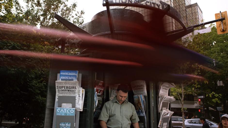 Supergirl flies past a newsstand on her way to save the city.