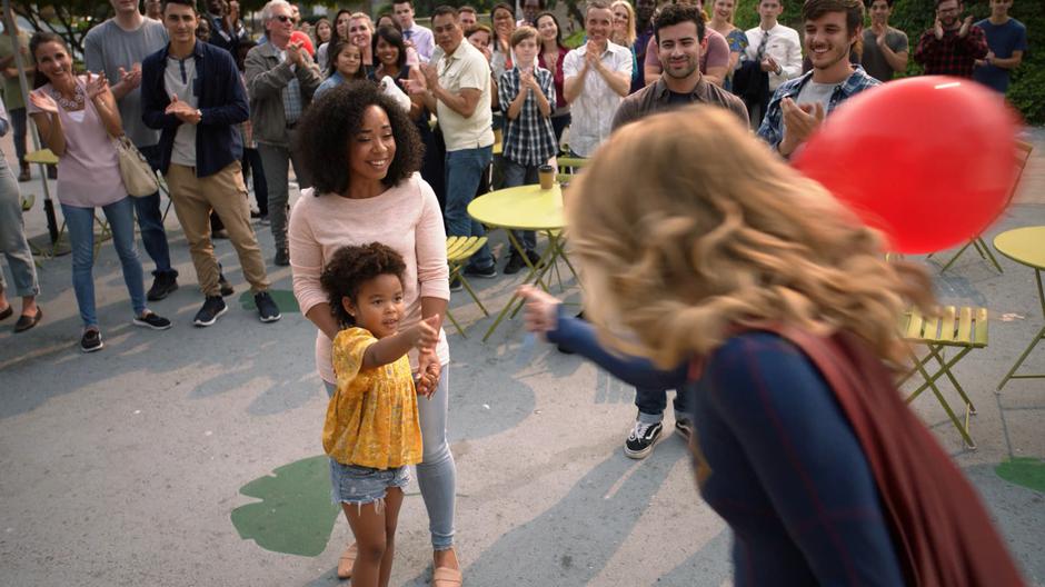 Kara hands the balloon back to the little girl while the girl's mother smiles at her.