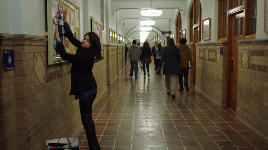 Mel finishes hanging up a poster while Harry walks past some students from down the hallway.