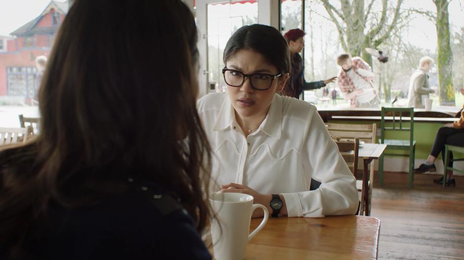 Mel looks over Niko's shoulder at the frozen scene outside the coffee shop window.