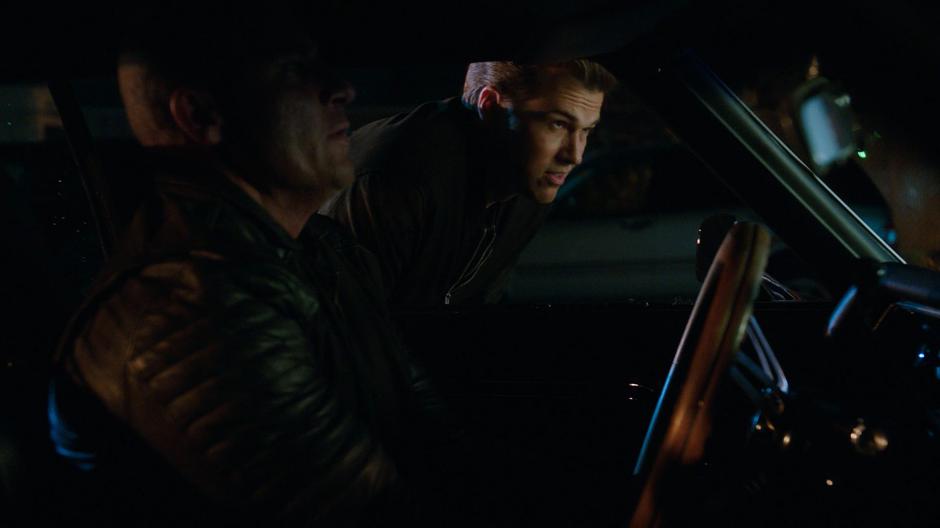 Nate leans down next to the missing window as Mick hotwires the car.