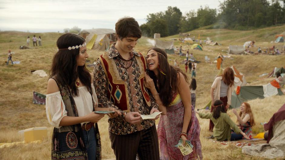 A hippie leans against Ray's shoulder after handing him and Zari fliers.