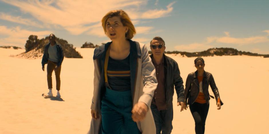 Ryan, the Doctor, Graham, and Yasmin walks across the hot sands.